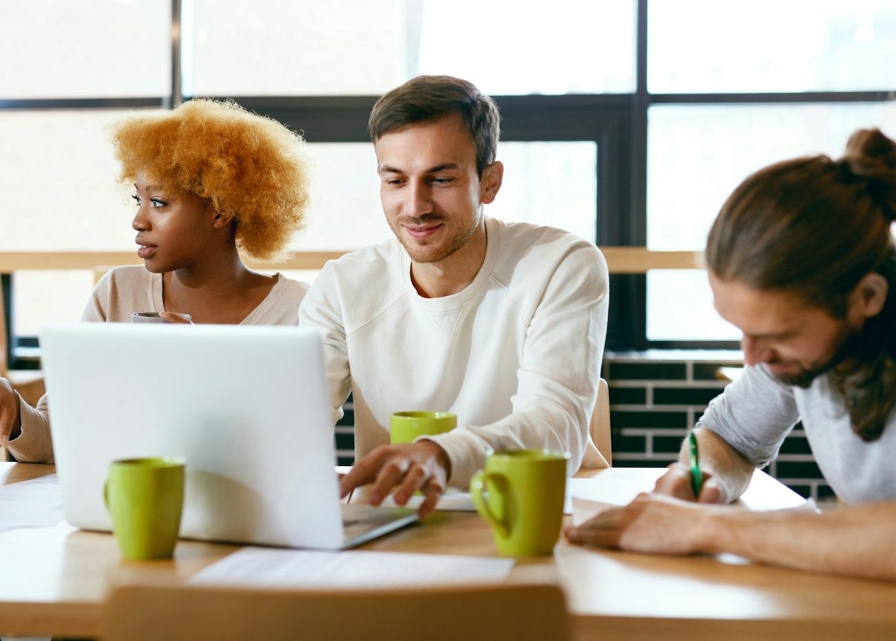Passgenaue Personalvermittlung Drei glückliche Personen arbeiten gemeinsam an einem Schreibtisch. Ein Mann blickt in sein Notebook. Drei Kaffeetassen sind zu sehen.